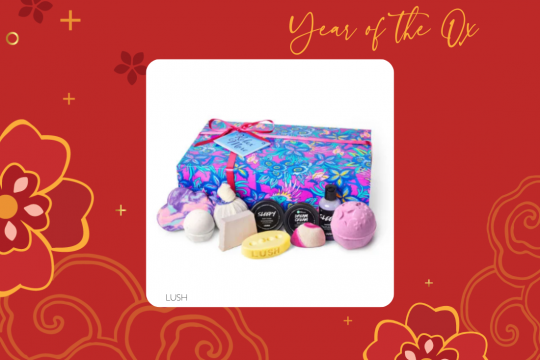 A white square frame at the center of a red floral background, with an image of a Lush bath bomb gift set. The text at the top says "Year of the Ox" and the text at the bottom says LUSH. 