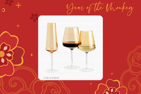 A white square frame at the center of a red floral background, with an image of three golden wine glasses - one for white wine, one for red, and one flute for champagne. The text at the top says "Year of the Monkey" and the text at the bottom says "Crate and Barrel." 